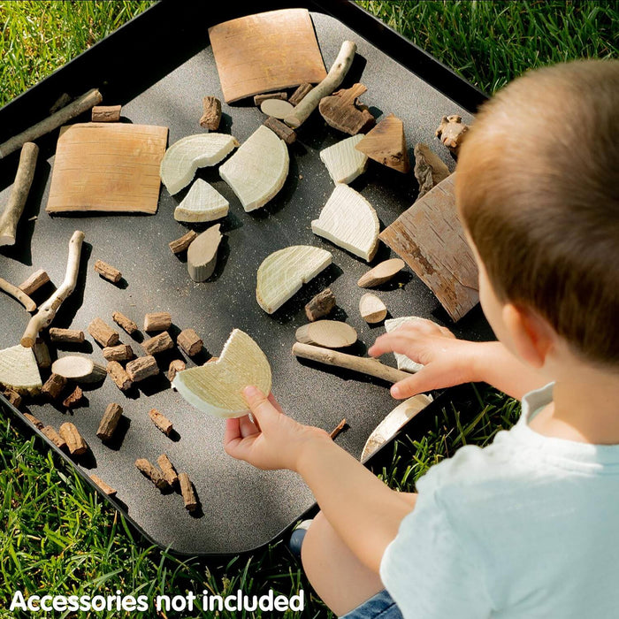 Child plays with natural wood pieces on a large square black tray with rounded corners, set on green grass.