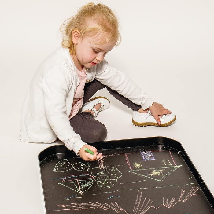 Child drawing with chalk on a large, shallow black rectangular tray with raised edges and rounded corners.