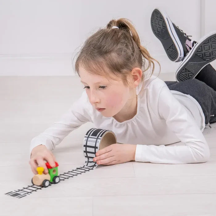 A child unrolling a roll of black and white railway track tape onto the floor for a small wooden toy train.