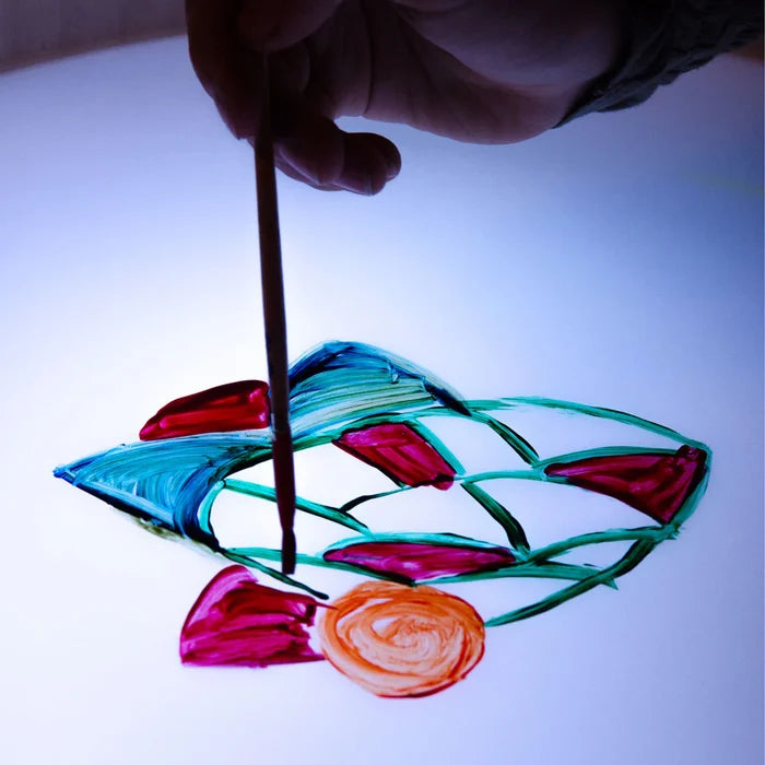Hand paints colorful abstract shapes on a bright circular light table surface with a paintbrush.