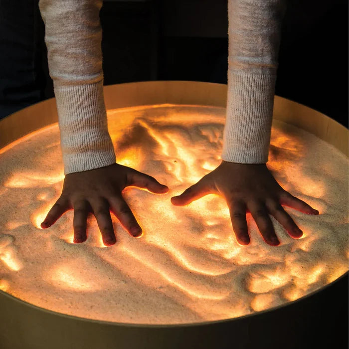 Hands pressing into sand on a round light table, with warm orange light glowing through the textured grains.
