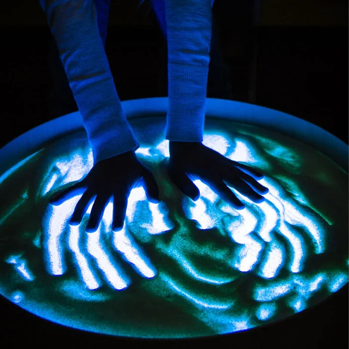 Child's hands creating patterns in sand on a round table illuminated with a bright cyan blue glow.