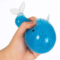 Hand squeezing a blue narwhal squishy filled with water beads, featuring a white tail and horn.