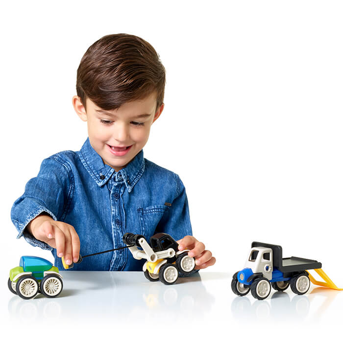 Boy in a denim shirt playing with a yellow crane truck, blue flatbed, and green toy car on a white background.