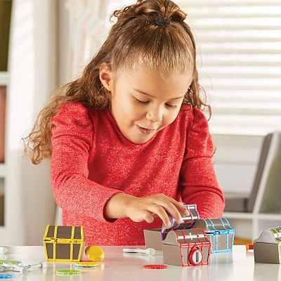 A young girl in a red shirt opens colorful plastic treasure chests surrounded by toy coins on a white table.