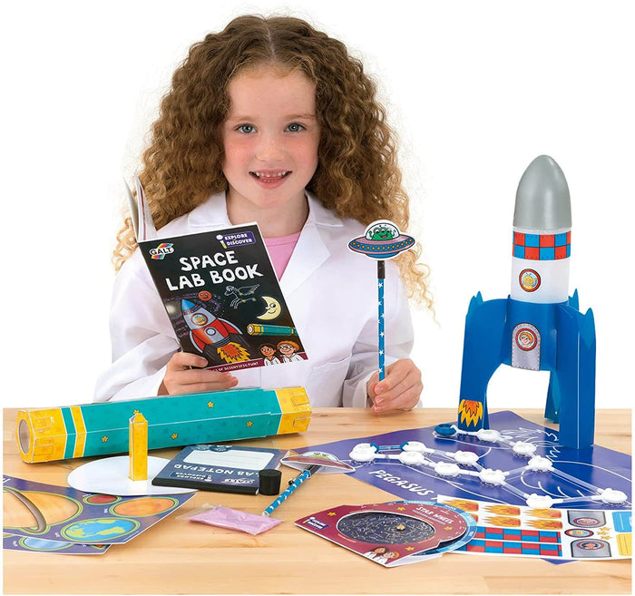 Girl in lab coat with space science kit: blue toy rocket, activity book, teal telescope, and star maps on a wooden table.