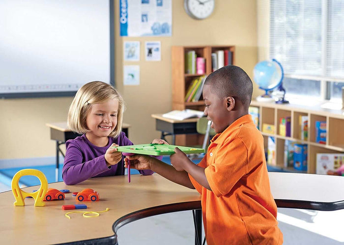 Two children play with a green magnetic tray, purple wand, and yellow horseshoe magnet at a classroom desk.