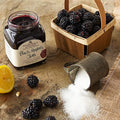 "Open jar of Stonewall Kitchen Black Raspberry Jam with a basket of fresh black raspberries, lemon wedge, and sugar on a rustic table."
