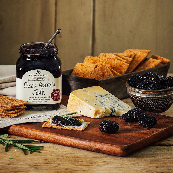 "Stonewall Kitchen Black Raspberry Jam served with crackers, wedge of blue cheese, and fresh black raspberries on a wooden board."