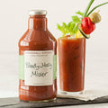 "Stonewall Kitchen Bloody Mary Mixer bottle with garnished drink – Glass bottle of Bloody Mary Mixer alongside a tall glass filled with the cocktail, garnished with celery, cucumber, pepper, and olive."