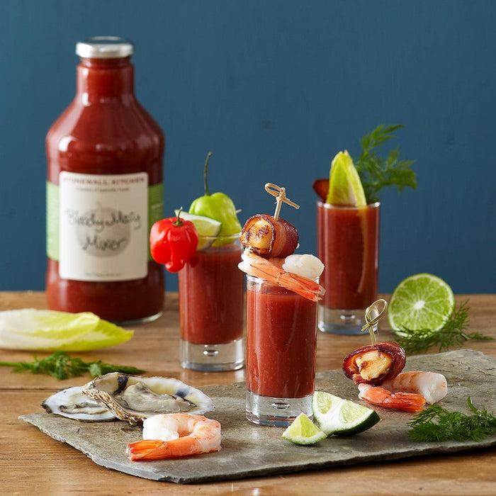 "Stonewall Kitchen Bloody Mary Mixer with shrimp and oyster garnishes – Bottle of Bloody Mary Mixer with three cocktail glasses, garnished with shrimp, bacon-wrapped dates, lime, and oysters."