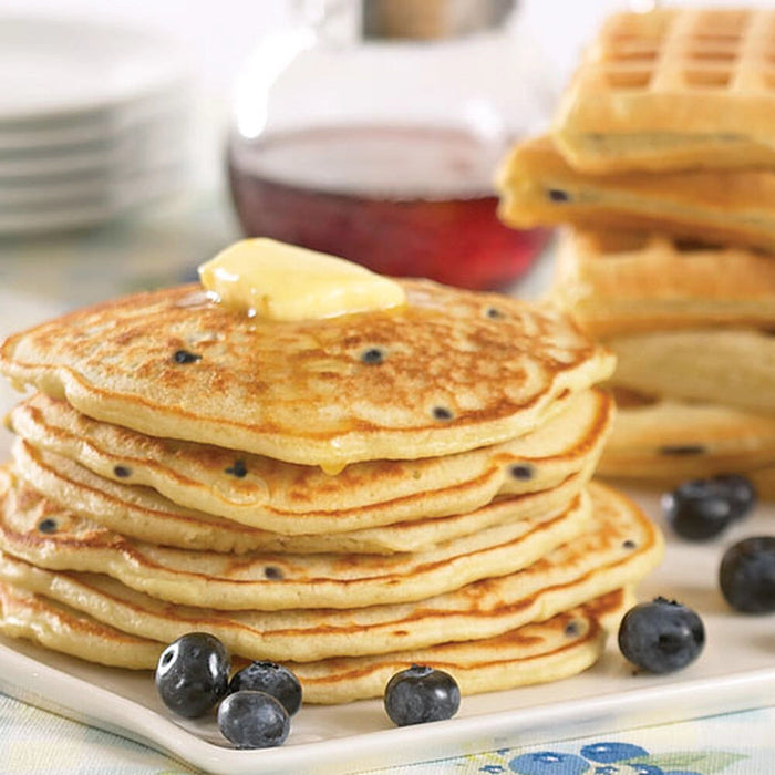 "Stack of fluffy blueberry pancakes topped with melting butter, fresh blueberries, and golden waffles in the background, served with maple syrup."