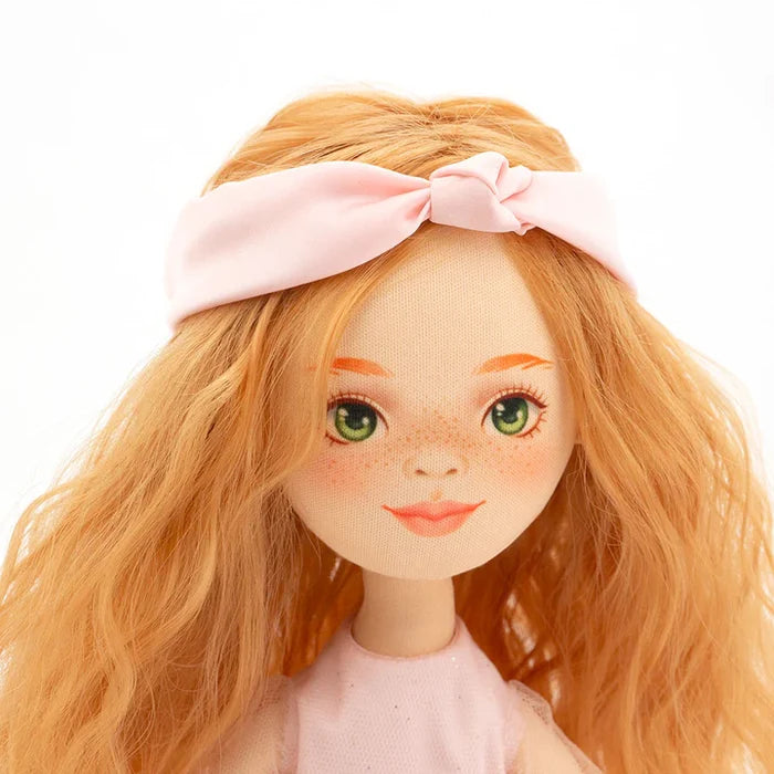 Doll with wavy orange hair, green eyes, and freckles wearing a light pink knotted headband and dress.