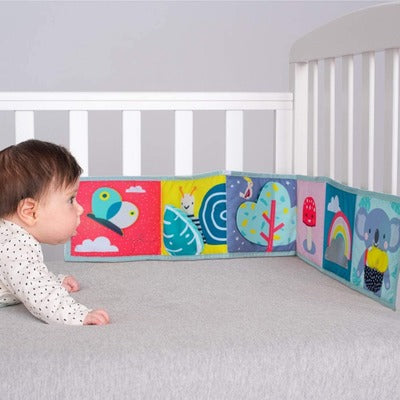 Baby in a crib looking at a colorful soft accordion book with animal illustrations like a butterfly and a koala.