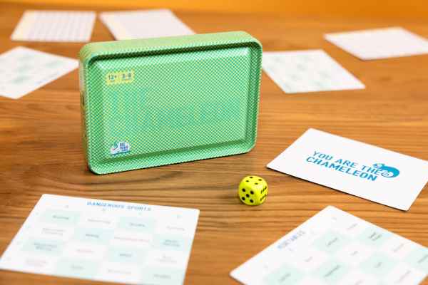 Green textured metal tin box, yellow die, and white game cards on a wooden table.