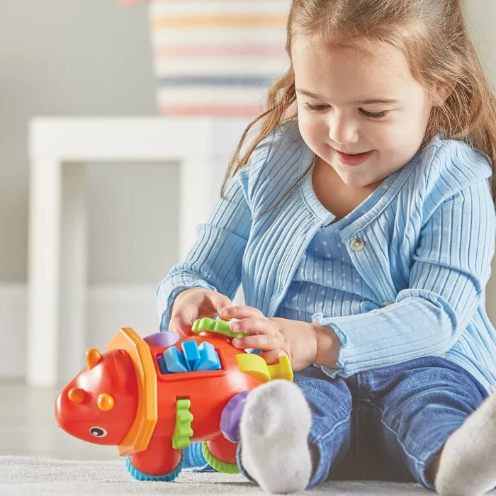 Smiling girl plays with a red triceratops fidget toy with colorful blue gears, green wheels, and orange details.