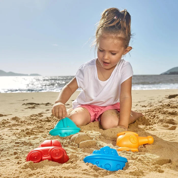 Child playing on a beach with red car, blue boat, teal boat, and orange helicopter sand molds.