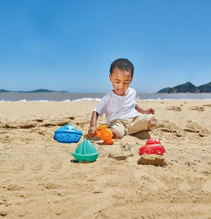 Young boy on beach with colorful sand molds shaped like a blue boat, green sailboat, red car, and orange airplane.