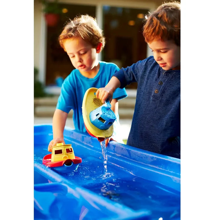 Two boys play with red, yellow, and blue toy tugboats in a blue water table, pouring water from a yellow boat.