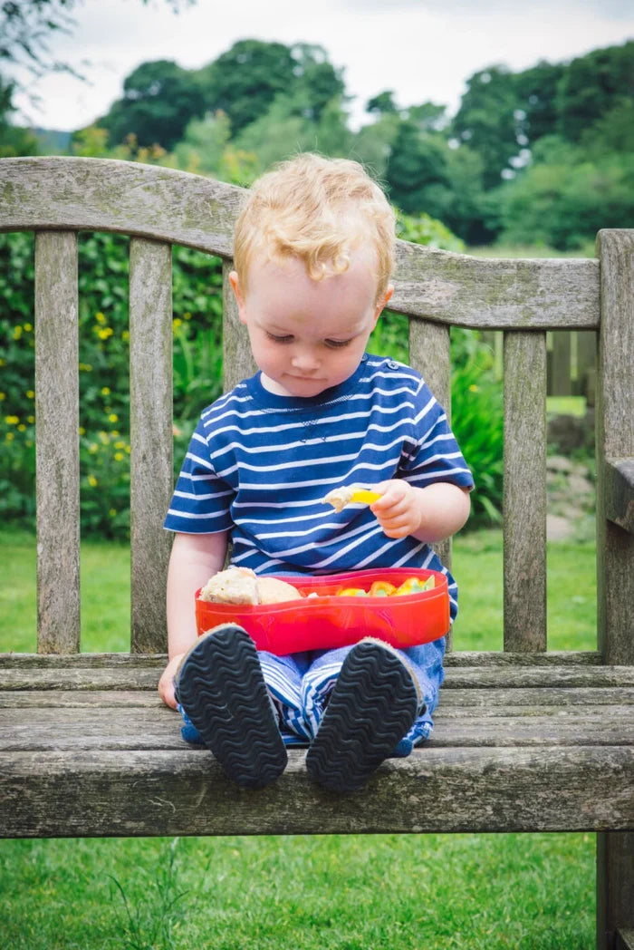 Toddler sitting on a wooden bench with a red heart-shaped lunch container featuring a small dipping pot separator.