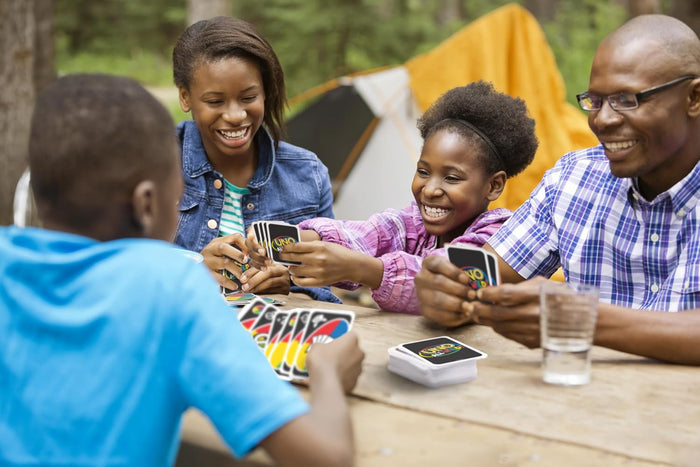 Family playing UNO All Wild at an outdoor table; black cards feature colorful rainbow-striped designs.