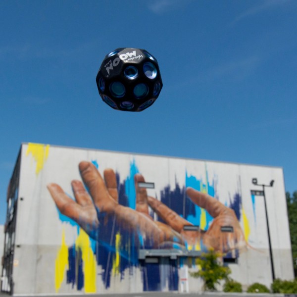 A black cratered Waboba Moon Ball with blue accents mid-air against a blue sky and an outdoor building mural.