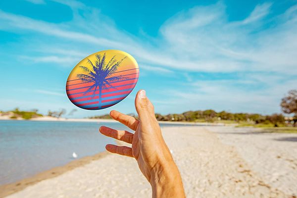 Hand catching a round yellow and blue flying disc with a palm tree graphic at a sunny beach under a blue sky.