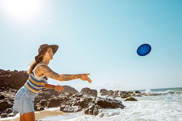 A woman throws a blue circular flying disc into the air at a sunny rocky beach with crashing ocean waves.
