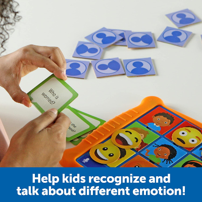Hands hold a green question card over an orange game board with diverse children's faces and yellow emoji expressions.