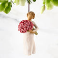 A Willow Tree style hanging ornament depicting a woman in a cream dress holding a bouquet of pink and red roses, suspended against a white marble background with green leaves hanging at the top.