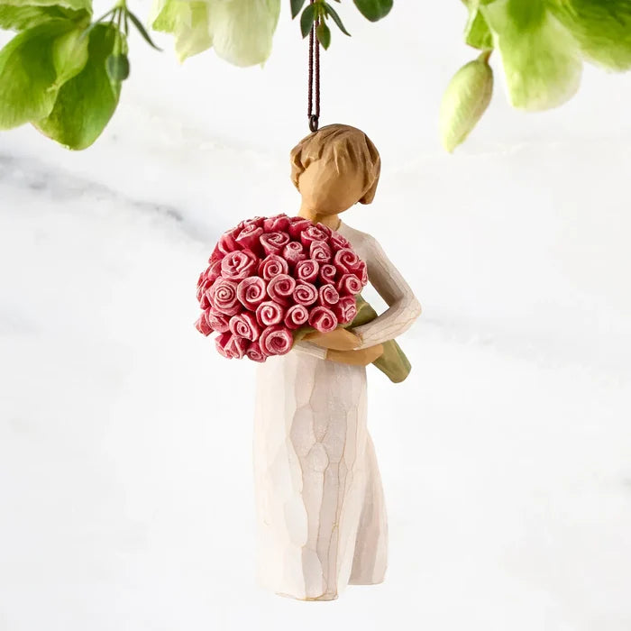 A Willow Tree style hanging ornament depicting a woman in a cream dress holding a bouquet of pink and red roses, suspended against a white marble background with green leaves hanging at the top.