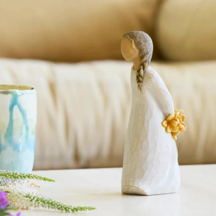 Willow tree figurine with yellow flowers behind her back on a table with a beige background