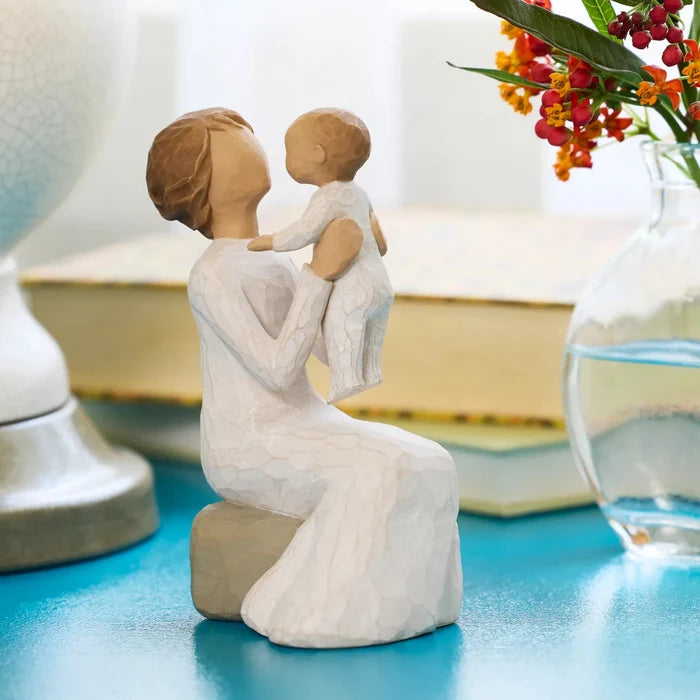 Willow Tree Grandmother figurine holding a baby o a blue table with a vase on the background.