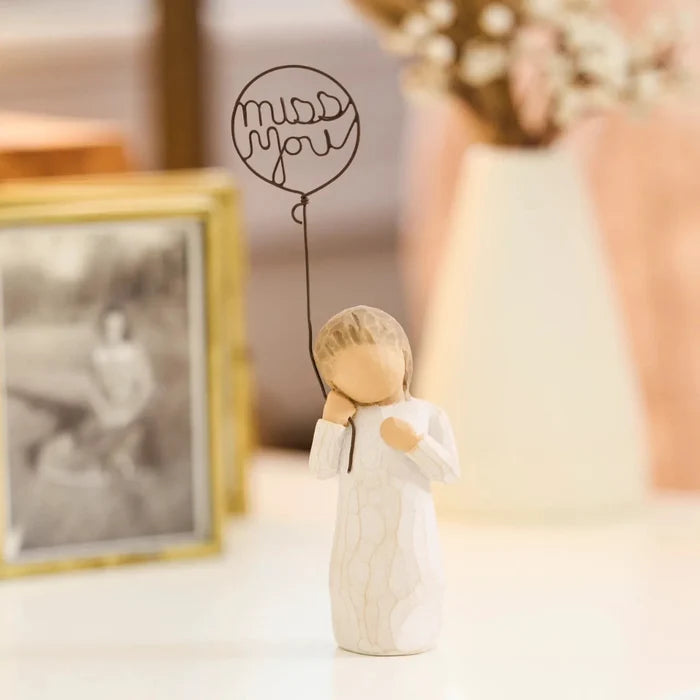 Small figurine of a person holding a balloon with 'miss you' text, next to a framed photo on a surface.